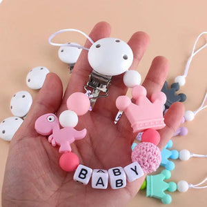 A hand gently holding the pink Little Dino Silicone Pacifier Chain, showcasing its secure Hemu pacifier clip and playful dinosaur design.